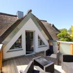Terrasse mit Sitzgruppe vor einem Haus mit Strohdach und Glasfront.