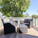 Terrasse mit Gartenmöbeln, Holzdielen und Glasgeländer unter Bäumen.
