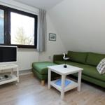 Ein Wohnzimmer mit grünem Sofa, weißem Couchtisch und Fernseher. Fenster mit Blick nach draußen.