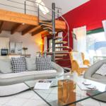 Wohnbereich mit Sofa, Sessel und Glas-Tisch. Hintergrund: Küche und Wendeltreppe.