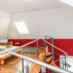 Schlafzimmer mit Dachschrägen, Holzboden und roter Wand. Bett, Treppe und Dachfenster sichtbar.