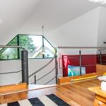 Dachgeschosszimmer mit Holzboden, Metallgeländer und großem Fenster. Bett und Kleiderständer sind sichtbar.