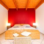 Schlafzimmer mit Holzbett, roter Wand und Holzdecke. Beleuchtung und Körbe sind vorhanden.