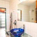 Badezimmer mit blauem Waschbecken, Spiegel und Dusche. Holzwand und gefärbte Tür sichtbar.