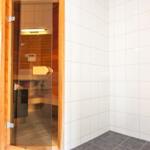 Sauna mit Holzwand und Glas Tür. Nebenraum mit weißen Fliesen und Dusche.