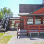 Rotes Ferienhaus mit Veranda, Treppe und Terrasse. Umgeben von Grün und Bäumen.