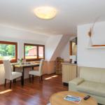 Wohn- und Essbereich mit Sofa, Tisch, Stühlen und Fenstern. Holzboden und weiße Wände.