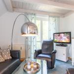 Wohnzimmer mit Sofa, Sessel, TV und Glas-Tisch unter Holzbalkendecke.