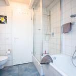 Badezimmer mit Dusche, Badewanne und Toilette. Weiße Fliesen und Holzbalken an der Decke.