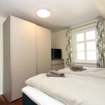 Schlafzimmer mit Doppelbett, Kleiderschrank, TV und Fenster mit Vorhängen.