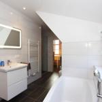 Badezimmer mit Badewanne, Waschbecken und Spiegel. Durchgang zur Sauna sichtbar.