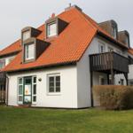 Wohnhaus mit rotem Ziegeldach, weißen Wänden und Balkonen. Grünflächen und Sträucher umgeben das Gebäude.