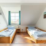 Zwei Einzelbetten mit blauen Bettwäschen, Holzrahmen und Fenster mit grünen Vorhängen.