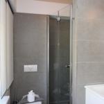 Bad mit Dusche, Waschbecken und Fenster. Graue Fliesen und weiße Sanitärgeräte.