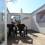 Terrasse mit Tisch, Stühlen und geschlossenem Sonnenschirm. Holzdecke und weiße Wände.
