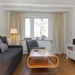 Wohnzimmer mit Sofa, Couchtisch und Fernseher. Fenster mit Vorhängen und Holzboden.