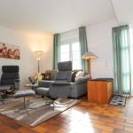 Wohnzimmer mit Sofa, zwei Sessel und Holzfußboden. Fenster mit grünen Vorhängen.