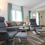 Wohnzimmer mit grauen Sesseln, Sofa und Tisch. Fenster mit grünen Vorhängen. Teppich und Holzboden.