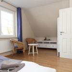 Ein Schlafzimmer mit Bett, Stuhl, Tisch und Fenster. Holzfußboden und weiße Wände.