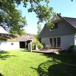 Haus mit Terrasse, Garten und Bäumen unter blauem Himmel.