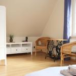 Zimmer mit Bett, Schrank, Tisch und zwei Stühlen. Fenster mit Vorhang. Holzboden.