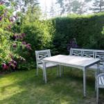 Helle Gartenmöbel auf Rasenfläche mit Blumenhecke und Bäumen im Hintergrund.