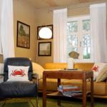 Ein Wohnzimmer mit gelbem Sofa, schwarzer Ledersessel und Holztisch. Fenster mit weißen Vorhängen.