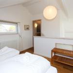 Schlafzimmer mit Bett, Fenster und Holzbank. Decke mit Balken und Pendelleuchte.
