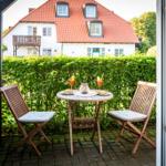 Terrasse mit Tisch und zwei Stühlen, dahinter grüner Heckenzaun und weißes Haus mit rotem Dach.
