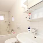 Badezimmer mit Badewanne, Dusche, Waschbecken und Toilette. Weiße Fliesen und Regale.