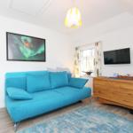 Ein Wohnzimmer mit blauem Sofa, Holzschrank und Wandbild. Fenster mit Vorhängen.