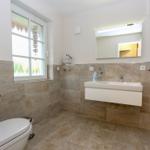 Badezimmer mit Toilette, Waschbecken und Dusche. Fenster mit Vorhang und Spiegel über Waschbecken.