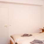 Schlafzimmer mit Bett, Nachttisch und Wandlampe. Holzschrank und Tür sind sichtbar.