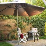 Terrasse mit Sonnenschirm, Tisch und zwei Stühlen. Hintergrund: Holzzaun und grüne Pflanzen.