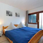 Schlafzimmer mit Holzbett, blauer Bettwäsche, Fenster mit Blick auf Wasser und Wald.