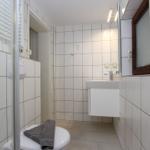 Badezimmer mit Dusche, Waschbecken und Toilette. Weiße Fliesen und Holzfenster.