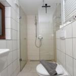 Badezimmer mit Dusche, Toilette und Waschbecken. Weiße Fliesen und Fenster mit Holzrahmen.
