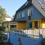 Ein zweistöckiges Haus mit blau-gelber Fassade, Dachgauben und einem blauen Tor. Fahrräder stehen vor dem Haus.