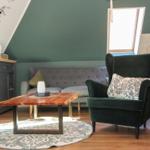 Ein Wohnbereich mit grünem Sessel, grauem Sofa und Holztisch. Dachfenster und Wandmalerei sichtbar.