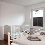 Zimmer mit Babybett, Bett und Fenster mit Jalousien.