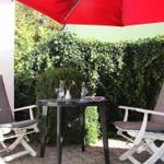 Terrasse mit Tisch, zwei Stühlen und rotem Sonnenschirm. Hintergrund: grüner Heckenzaun.