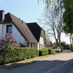 Wohnhaus mit dunklem Dach und grüner Hecke, umgeben von Bäumen und blühenden Sträuchern.