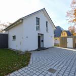 Moderne weiße Villa mit gepflastertem Hof und grünem Garten. Umgeben von Bäumen mit Herbstlaub.