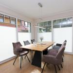 Esszimmer mit Holztisch, Stühlen und großen Fenstern mit Jalousien.