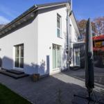 Moderne weiße Villa mit Terrasse und Garten. Schwarze Dachrinne und Fenster sind sichtbar.