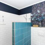 Bad mit Dusche, blauen Fliesen und floraler Tapete. Holztür und Pflanzen sind sichtbar.