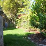 Garten mit Rasen, Sträuchern und Schild. Im Hintergrund ist ein Gartenhaus sichtbar.