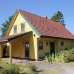Gelbes Haus mit rotem Dach und grünen Fensterrahmen, umgeben von Grün und Blumen.