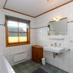Badezimmer mit Badewanne, Waschbecken und Holzschrank. Fenster mit Jalousie und Heizkörper.