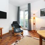 Wohnzimmer mit Fernseher, Sessel, Tisch und Fenster. Holzboden und Wandbild.